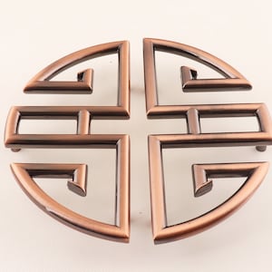 Può includere: Un sottobicchiere rotondo decorativo in metallo color rame con un design geometrico. Il design è un simbolo cinese stilizzato con una forma circolare e un motivo a croce.