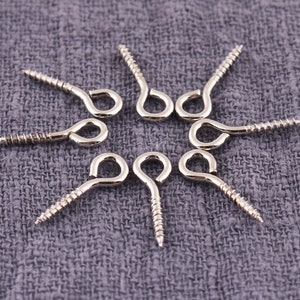May include: A close-up of eight silver screw-in hooks with a loop at the end. The hooks are arranged in a circle on a gray fabric background.
