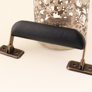 May include: A black leather handle with antique brass hardware. The handle is attached to two rectangular plates with two holes each.