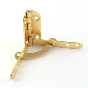 Puede incluir: Bisagra de esquina de metal dorado con un brazo redondeado y curvo. La bisagra tiene una serie de pequeños agujeros para tornillos.