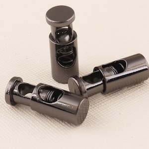 Puede incluir: Tres topes de cordón de metal negro con un mecanismo de resorte. Los topes son cilíndricos y tienen un acabado liso y brillante.