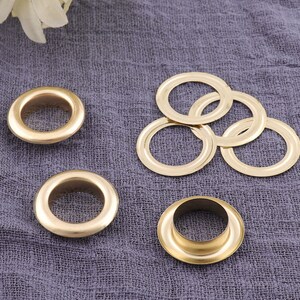 Könnte beinhalten: Ein Satz goldfarbener Metallstifte für Bastel- und Nähprojekte. Die Ösen sind in verschiedenen Größen und auf einem grauen Stoffhintergrund angeordnet.