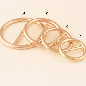May include: Four gold-colored metal rings with a bar across the center, in varying sizes. The rings are arranged diagonally, with the largest ring at the top left and the smallest at the bottom right. The rings have a shiny, reflective surface.