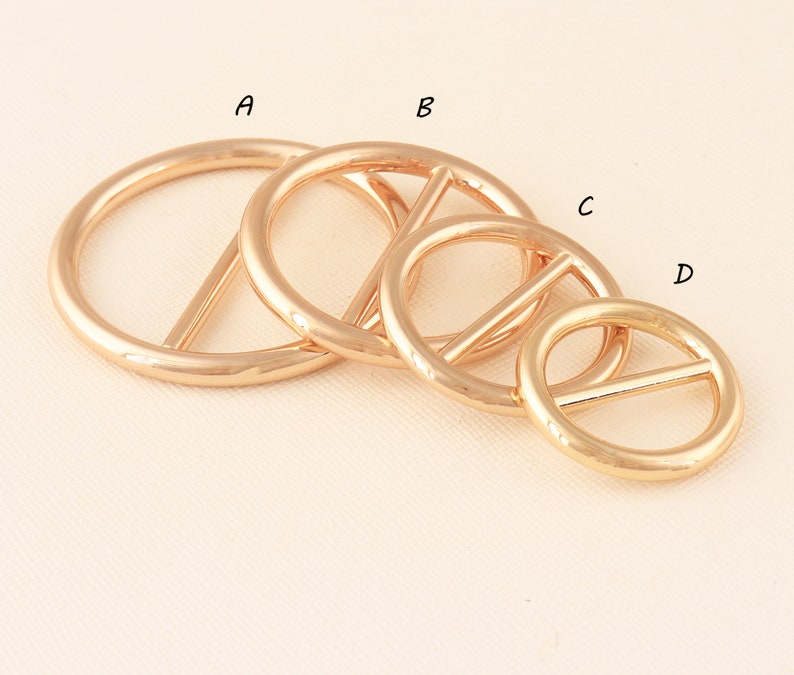 May include: Four gold-colored metal rings with a bar across the center, in varying sizes. The rings are arranged diagonally, with the largest ring at the top left and the smallest at the bottom right. The rings have a shiny, reflective surface.