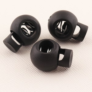 Puede incluir: Tres topes de plástico negro con un resorte metálico en el interior. Los topes tienen una forma redonda con un pequeño agujero en el centro y una abertura lateral para sujetar a un cordón o cuerda.