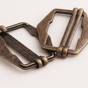 Puede incluir: Dos hebillas deslizantes de metal de color dorado antiguo. Las hebillas son de forma hexagonal y tienen un acabado desgastado y envejecido.