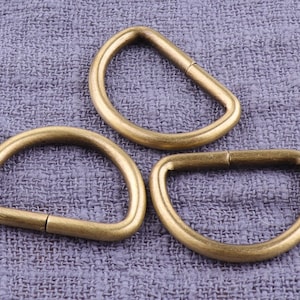 May include: Three gold-toned metal D-rings of varying sizes. The rings are smooth and have a slightly rounded edge.