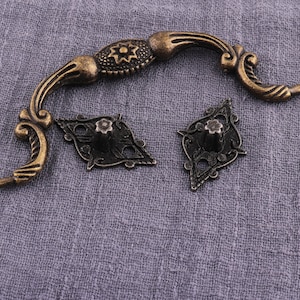 May include: Antique gold-toned cabinet hardware with a decorative design. The handle is shaped like a curved arch with a floral design. The two matching knobs are diamond-shaped with a floral design.