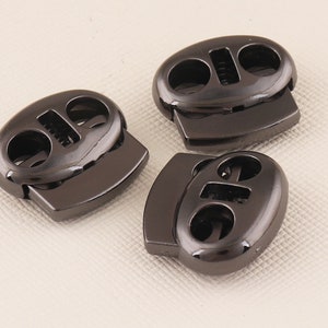 Puede incluir: Tres cierres de cordón de plástico negro con acabado brillante. Cada cierre tiene dos agujeros para enhebrar el cordón.