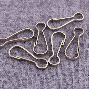May include: A collection of silver-colored metal swivel clasps, also known as snap hooks, arranged on a gray fabric surface. The clasps have a spring-loaded gate and a rounded loop at one end.