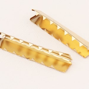 May include: Two gold-colored metal picture frame clips. Each clip is rectangular with a series of triangular cutouts along one edge. The clips have a shiny, reflective surface and are designed to secure the back of a picture frame.