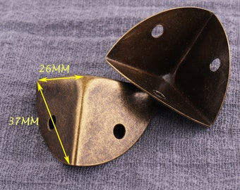 May include: Two antique gold corner protectors, each measuring 37mm by 26mm. The protectors are made of metal and have a distressed finish.