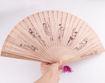 Puede incluir: Un abanico plegable de madera con un borla rosa. El abanico está decorado con un delicado patrón de puntos blancos y un diseño de mujeres con ropa tradicional.