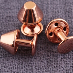 Puede incluir: Tres remaches de metal de color cobre con una parte superior abovedada y una base redonda. Los remaches tienen un poste que se extiende desde la base.