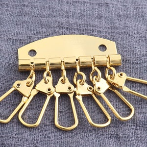 May include: Gold-toned metal key chain with six key rings. The key chain has a rectangular base with a rounded top and a hinged clasp.