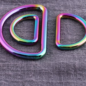 May include: Two iridescent metal D-rings, one larger than the other, are shown against a gray fabric background.