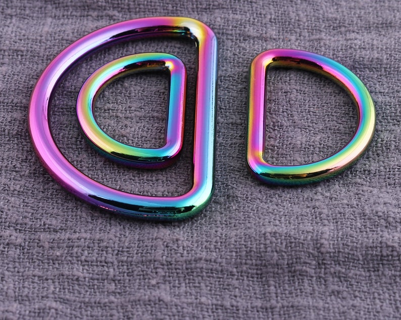 May include: Two iridescent metal D-rings, one larger than the other, are shown against a gray fabric background.