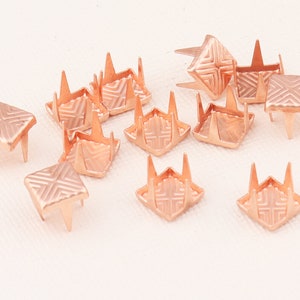 Peut inclure: Un groupe de 12 clous pyramidaux en métal rose gold avec un motif géométrique. Les clous ont quatre pointes sur le dessous.