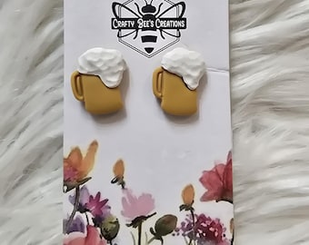 May include: A pair of novelty earrings shaped like mugs of beer. The earrings are yellow with white foam and are displayed on a card with the text "Crafty Bee's Creations" and a floral design.