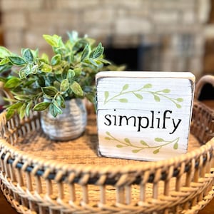 May include: A decorative wooden block with the word "simplify" in black lettering, accented by green leaf designs. The block sits on a woven tray with a small potted plant. The tray is made of natural materials.