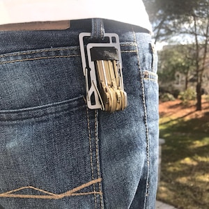 May include: A silver key organizer clipped to the back pocket of blue jeans. The key holder is rectangular with a black elastic band holding several keys. The jeans are a medium wash denim.