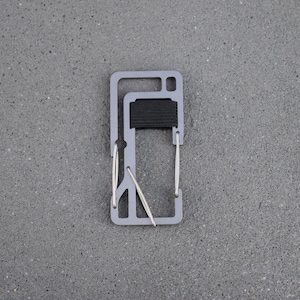 May include: A silver-colored multi-tool with a black rubber band attached to the top. The tool has a variety of features, including a bottle opener, a pry bar, and a wire cutter.