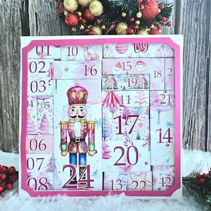May include: A pink and white Christmas advent calendar with a decorative pink frame. The calendar features numbered blocks with festive illustrations, including a nutcracker, Christmas trees, and snowflakes. The numbers range from 1 to 24.