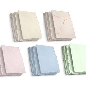 Pode incluir: Cinco pilhas de papel artesanal em creme, bege claro com inclusões, rosa pálido, azul claro e verde claro. Cada pilha tem uma borda irregular, dando um toque rústico e artesanal.