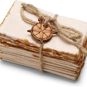 K&ouml;nnte beinhalten: Ein Stapel handgemachtes Papier, gebunden mit Schnur und einem Holz-Kompass-Anh&auml;nger. Das Papier hat ein nat&uuml;rliches, rustikales Aussehen und Gef&uuml;hl.