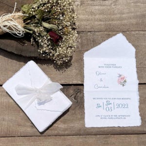 Puede incluir: Invitaci&oacute;n de boda en papel hecho a mano con textura r&uacute;stica. La invitaci&oacute;n presenta un dise&ntilde;o floral con una rosa rosa y texto azul. El texto dice "Juntos con sus familias Oliver & Camelia Les invitamos a unirse a nuestra boda Sun 03 2022 La una de la tarde Hotel Name-Londres, Reino Unido."