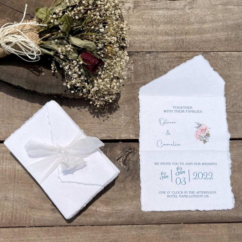 Puede incluir: Invitaci&oacute;n de boda en papel hecho a mano con textura r&uacute;stica. La invitaci&oacute;n presenta un dise&ntilde;o floral con una rosa rosa y texto azul. El texto dice "Juntos con sus familias Oliver & Camelia Les invitamos a unirse a nuestra boda Sun 03 2022 La una de la tarde Hotel Name-Londres, Reino Unido."