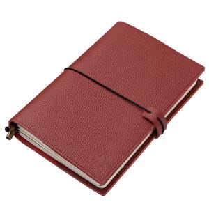 May include: A burgundy leather journal with a textured cover and a black elastic closure. The notebook is closed with a strap and a brass-colored metal detail. The pages are cream-colored, and a compass rose is embossed on the front cover.