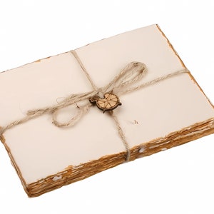 K&ouml;nnte beinhalten: Ein Stapel leeres, cremefarbenes Papier ist mit Schnur und einem Holz-Kompass-Anh&auml;nger zusammengebunden. Das Papier hat einen leicht gealterten, rustikalen Look.