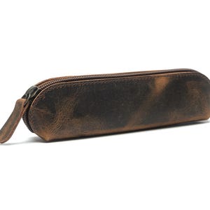 May include: A brown leather pencil case with a zipper closure. The case has a distressed finish, giving it a vintage look. The zipper pull is made of the same leather. This case is designed to hold pens, pencils, and other small stationery items.