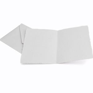 May include: A blank, white greeting card with a textured surface, open to display the inside pages. A matching white envelope is positioned to the left. The card and envelope are set against a white background.