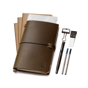 May include: A brown leather journal with a compass rose design, a black elastic closure, and a pen. Includes a metal clip, a bookmark, and two pen refills. The journal is accompanied by several kraft paper notebooks.