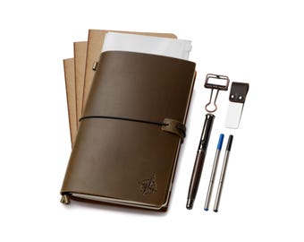 Könnte beinhalten: Ein braunes Lederjournal mit Kompassrosen-Design, schwarzem Gummibandverschluss und Stift. Enthält einen Metallclip, einen Lesezeichen und zwei Stiftminen. Das Journal wird von mehreren Notizbüchern aus Kraftpapier begleitet.