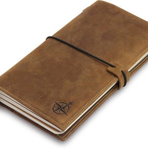 May include: A brown leather journal with a compass rose design. The journal is closed with a black elastic band and a leather strap. The pages are white with a cream-colored border. The journal is approximately 8 inches long and 5 inches wide.