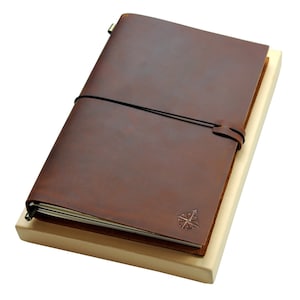 May include: A brown leather journal with a compass design on the cover. The journal is closed and has a black elastic band around it. The journal is sitting on a wooden surface.