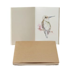 Op de afbeelding: Een set van drie kleine notitieboekjes. Eén notitieboekje is open en toont een aquarelillustratie van een kolibrie op een tak met roze bloemen. De andere twee notitieboekjes zijn gesloten, met een bruine kaft.