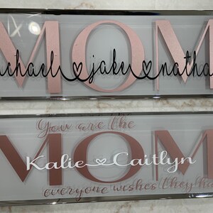Könnte beinhalten: Zwei gerahmte Glasschilder mit dem Wort "MOM" in großen rosa Buchstaben. Das obere Schild trägt die Namen "michael, jake, nathan" in schwarzer Schrift unter dem Wort "MOM". Das untere Schild trägt den Text "You are the MOM" in rosa Schrift mit den Namen "Kallie & Caitlyn" in weißer Schrift unter dem Wort "MOM". Das untere Schild trägt auch den Text "everyone wishes they had" in rosa Schrift unter den Namen.