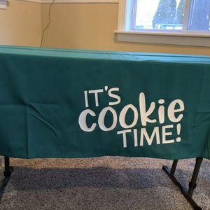 May include: A dark teal tablecloth with the phrase "IT'S cookie TIME!" in white lettering. The tablecloth is draped over a black metal folding table, suitable for events or displays.
