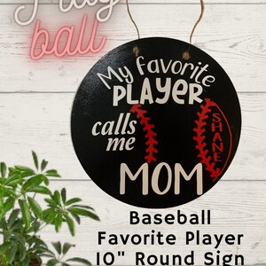 Op de afbeelding: Een rond, zwart houten bord met witte vinylbelettering met de tekst "Mijn favoriete speler noemt me MAMMA" met een rode baseballgrafiek en de naam "SHANE" in witte letters. Het bord heeft een diameter van 25,4 cm en wordt gepersonaliseerd met de naam van je favoriete speler. Rond bord met favoriete baseballspeler, 25,4 cm.