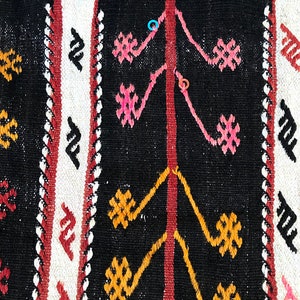 May include: Close-up of a black and white woven rug with intricate geometric patterns in red, pink, and yellow. The rug has a traditional design and is made of natural fibres.