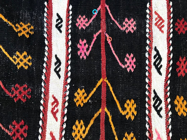 May include: Close-up of a black and white woven rug with intricate geometric patterns in red, pink, and yellow. The rug has a traditional design and is made of natural fibres.