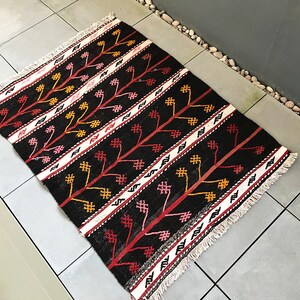 May include: A black, red and white patterned rug with a fringed border. The rug features a geometric design with stylised trees and flowers.