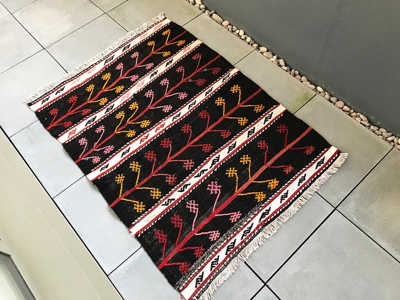 May include: A black, red and white patterned rug with a fringed border. The rug features a geometric design with stylised trees and flowers.