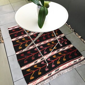 May include: A black and red patterned rug with white fringe on a patio with a white folding table and a potted plant.