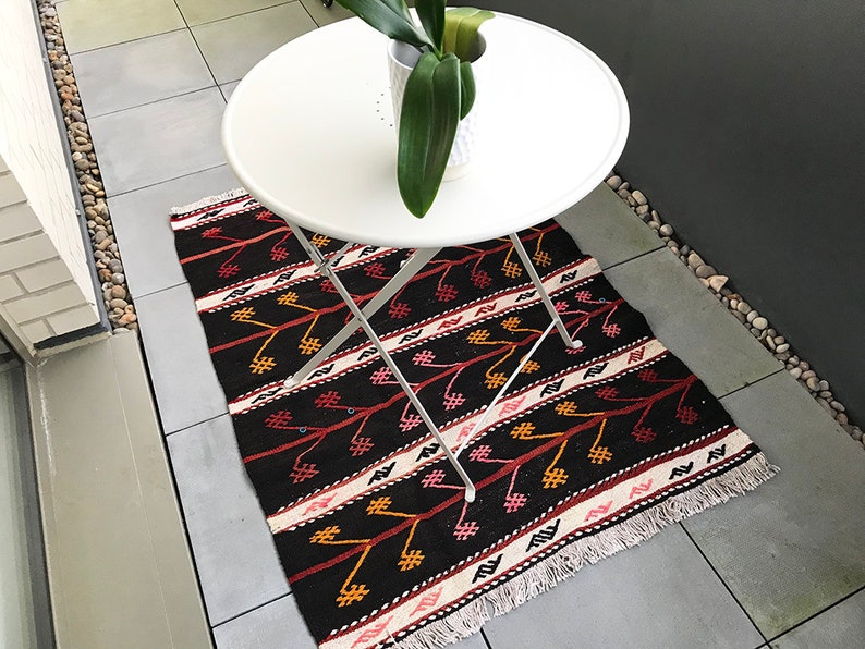 May include: A black and red patterned rug with white fringe on a patio with a white folding table and a potted plant.