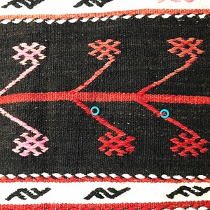 May include: Close-up of a black and red woven rug with a geometric pattern. The rug has a white border with a repeating pattern of stylised waves.